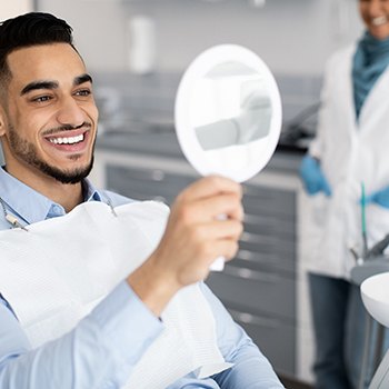 Man in blue shirt in dental chair smiling at treatment results in mirror
