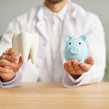 Dentist wearing white holding large model tooth in one hand and blue piggy bank in the other
